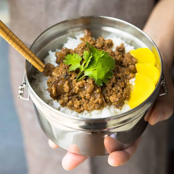 A stainless steel lunch tin of braised minced pork over rice, topped with coriander and pickled daikon, held in both hands.